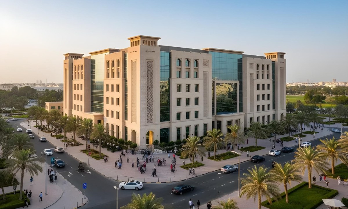 The image shows a modern building with glass windows, surrounded by palm trees, and people walking in front of it. Cars are on the road, and the scene is set in a sunny, urban environment.