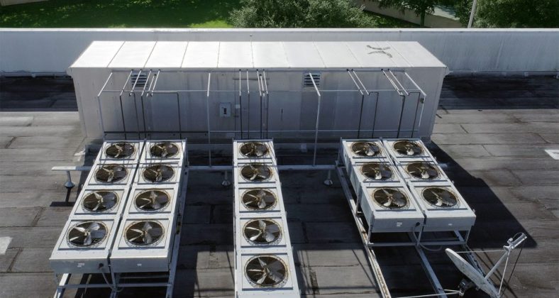 Top view of industrial air conditioning units installed on a flat rooftop, with a background of green trees and a clear sky